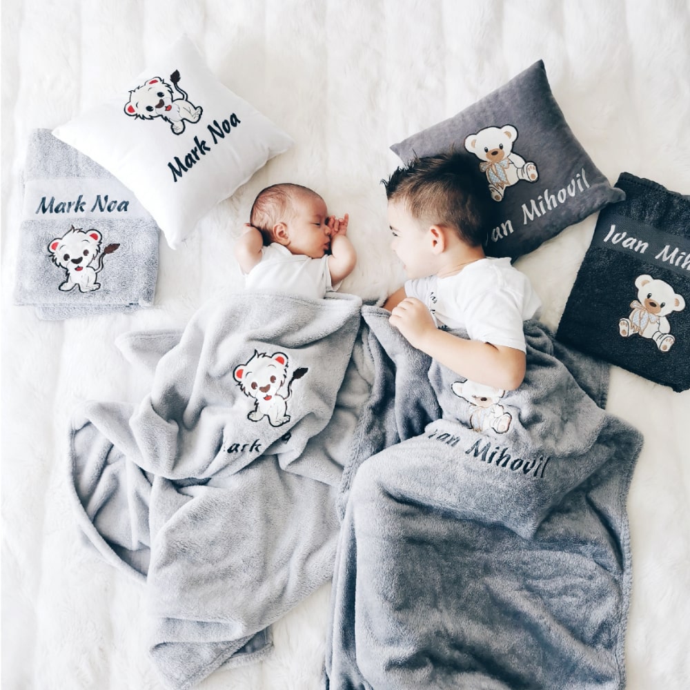 Schattige babyjongen geniet van zijn gepersonaliseerde set bestaande uit een kussen, deken en handdoek, allemaal aangepast met zijn naam. Naast hem geniet zijn oudere broer ook van dezelfde set.